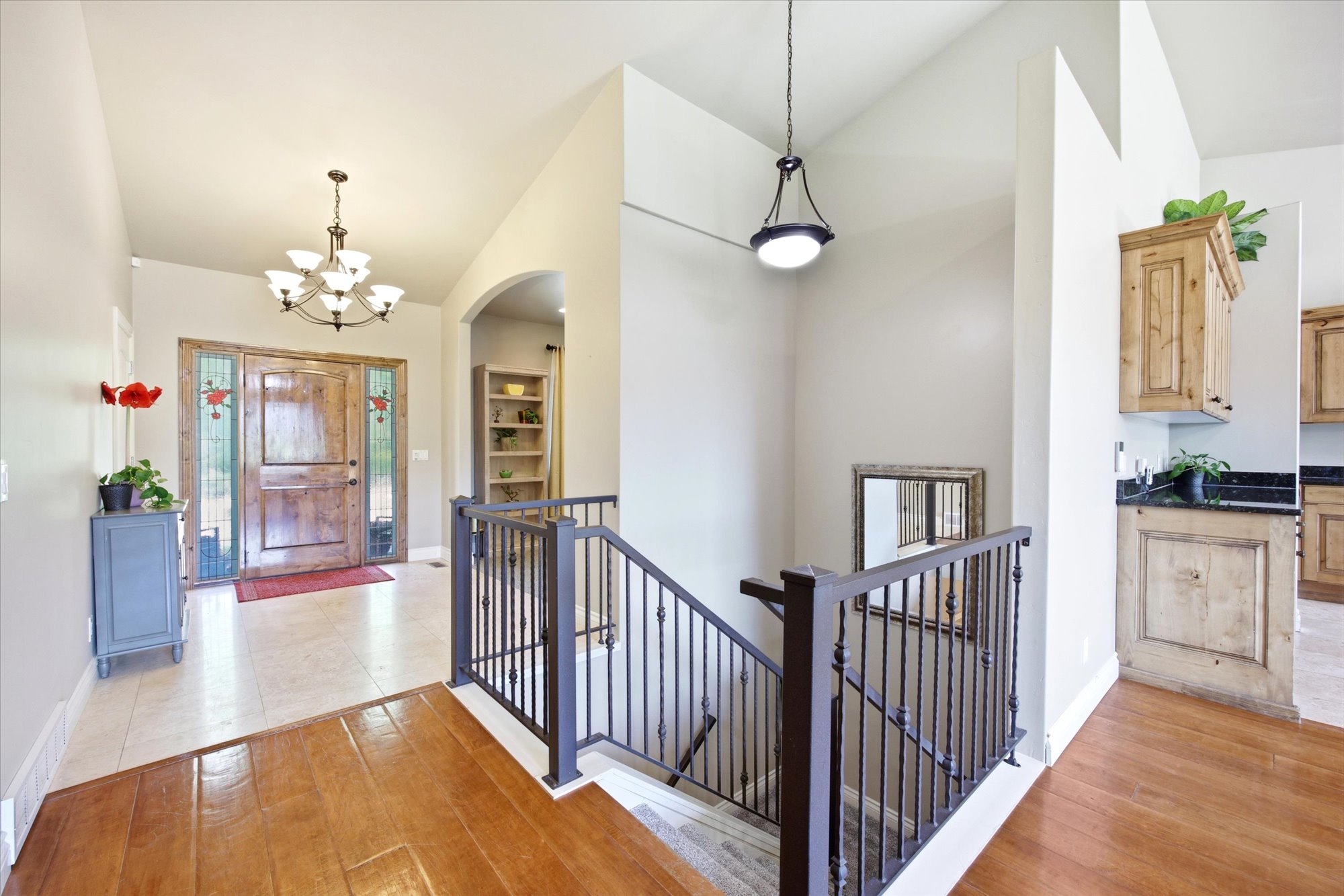 Foyer with wood-finish flooring transitioning to tile, featuring a rustic wooden entry door with sidelights, an ornate chandelier, and an open staircase with wrought iron balusters