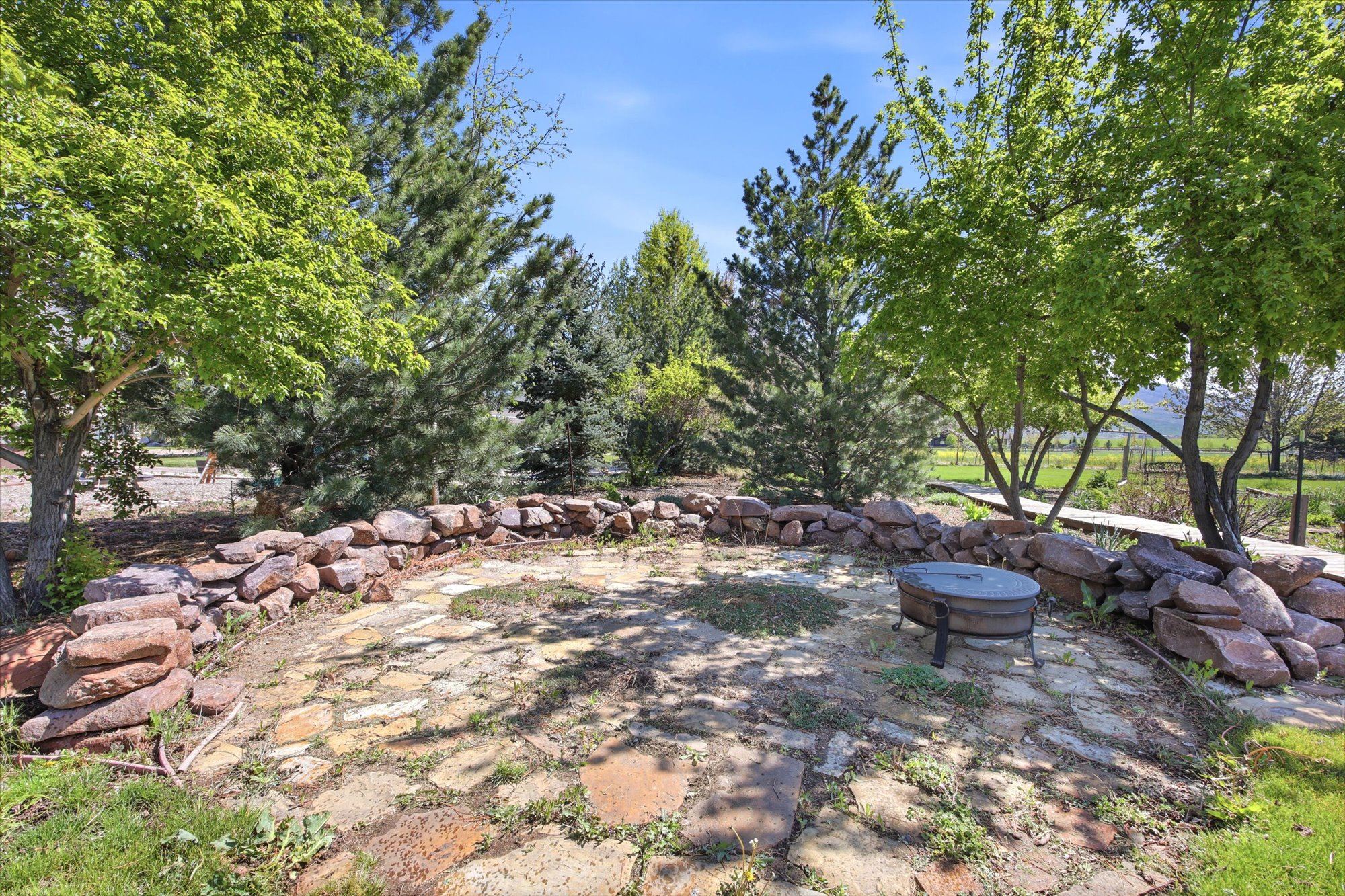 Outdoor gathering area featuring a stone-paved patio, a circular stone retaining wall, and mature trees providing natural shade