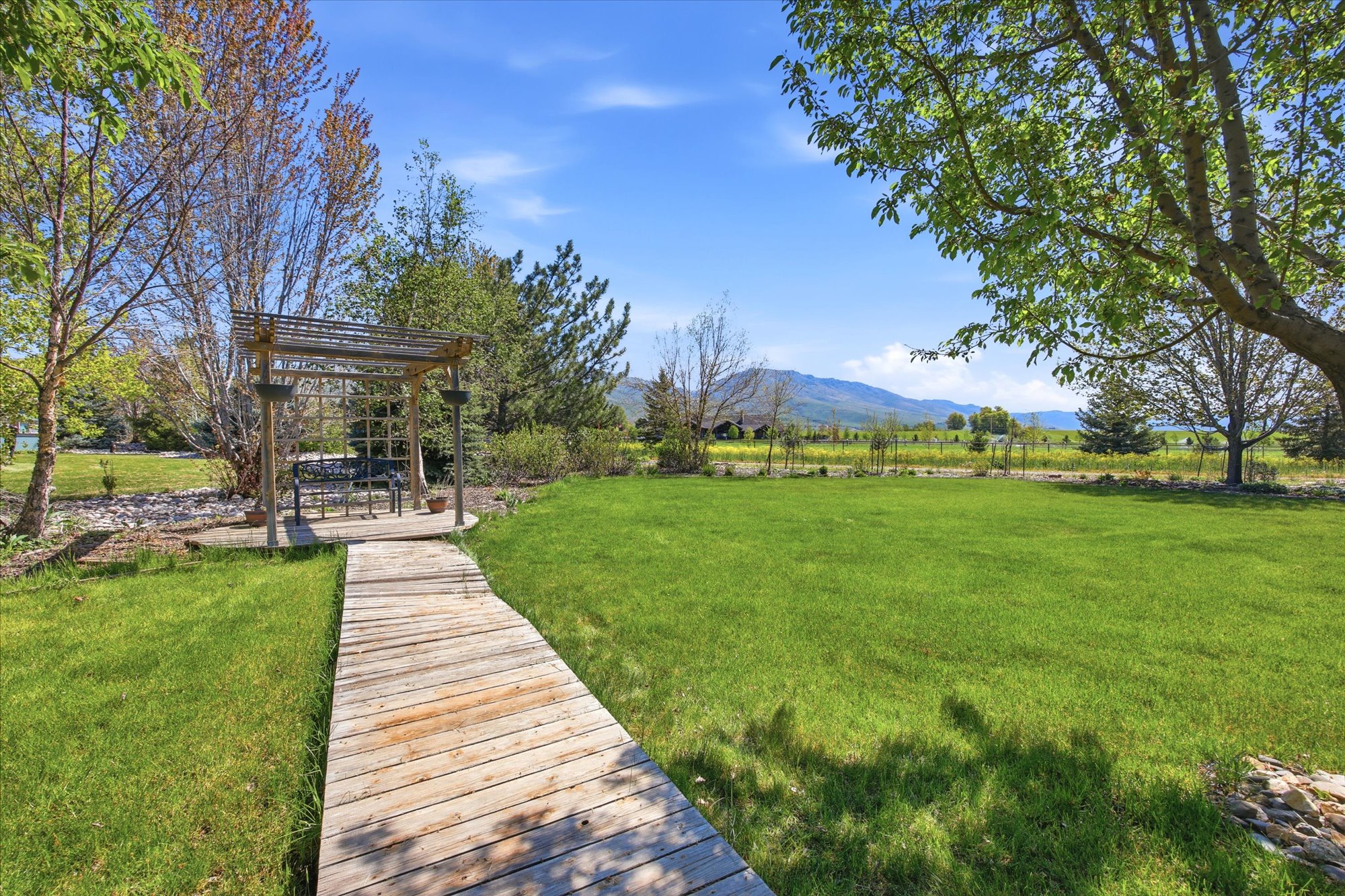 Expansive lawn featuring a wooden pergola with a seating area, bordered by mature trees and a wooden walkway