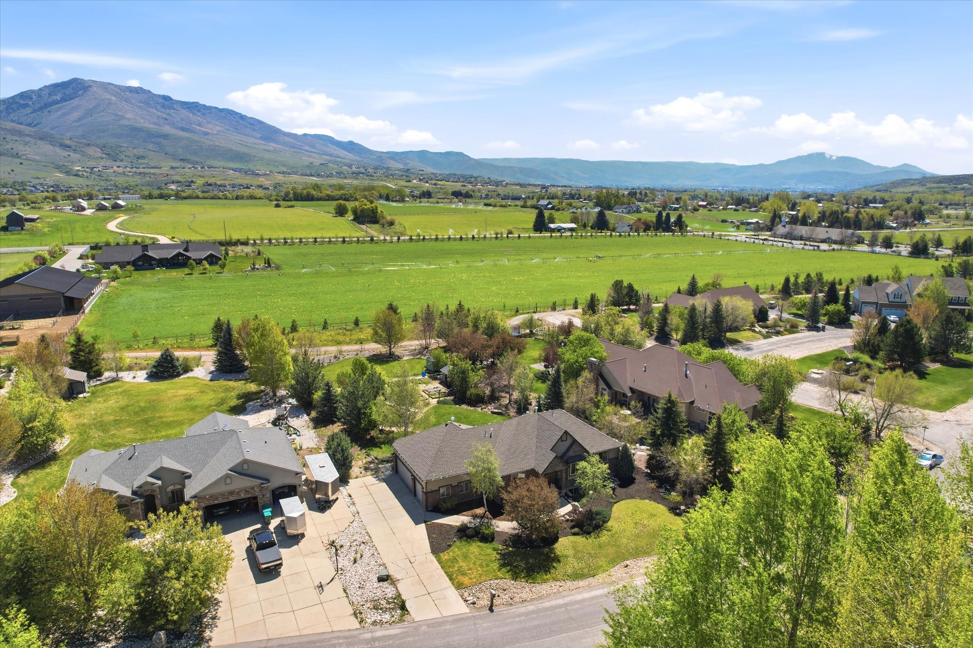 Expansive aerial perspective showcasing a residential property with a multi-car driveway, surrounding mature trees, and a spacious green lawn