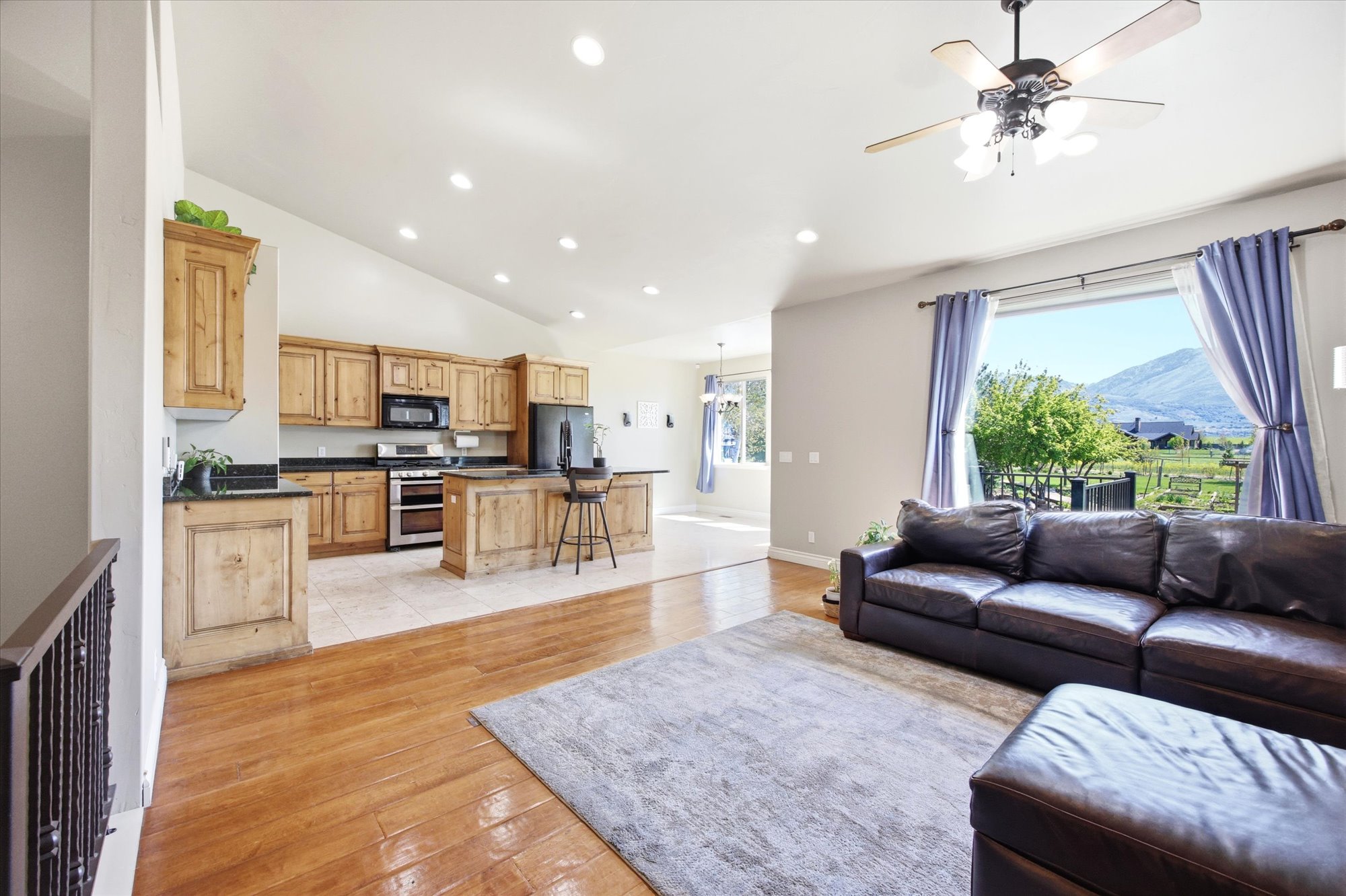 Open-concept living space featuring wood-finish flooring, a kitchen with natural wood cabinetry and dark countertops, recessed lighting, and a window framing a mountainous landscape