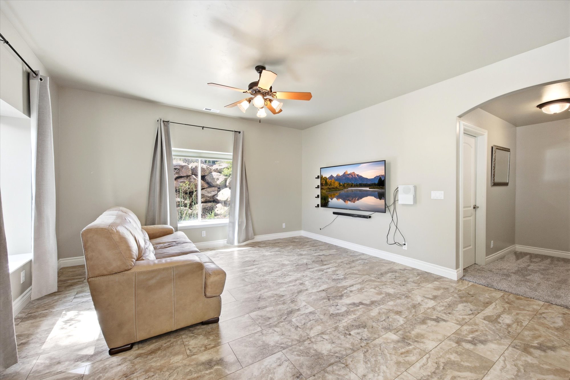 Open-concept living area featuring broad windows with natural light, large format tile flooring, a ceiling fan, and an arched entryway
