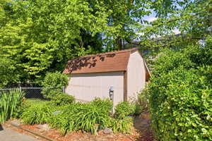 Gable-roofed utility shed with vertical siding, surrounded by mature trees and varied landscaping