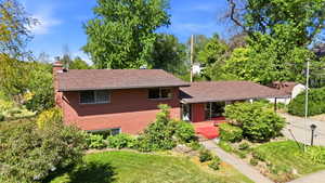 Brick exterior property featuring a covered entry, established landscaping, mature trees, a driveway, and a front lawn
