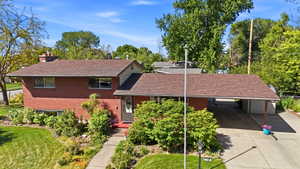 Brick exterior with a brown shingle roof, featuring a covered carport, mature landscaping, and a concrete driveway