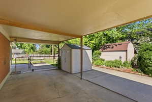 Covered concrete patio with a storage shed, brick exterior wall, chain-link fence, and landscaped perimeter