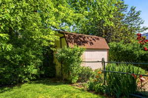Gable-roof storage shed with light-colored siding, surrounded by mature trees and varied plantings