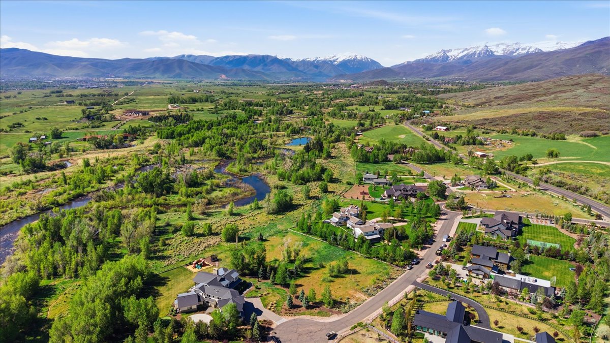 Expansive rural landscape featuring a winding river, mature trees, open fields, and distant snow-capped mountains under a clear sky