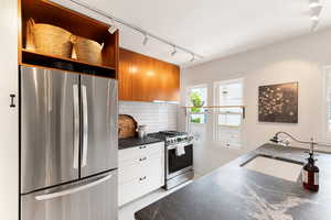 Modern kitchen featuring stainless steel appliances, dark stone countertops, wood-finish cabinetry, white subway tile backsplash, and track lighting