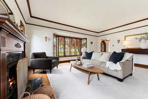 Living area with a bay window, hardwood flooring, a fireplace featuring a stone surround, decorative crown molding, and wall sconces