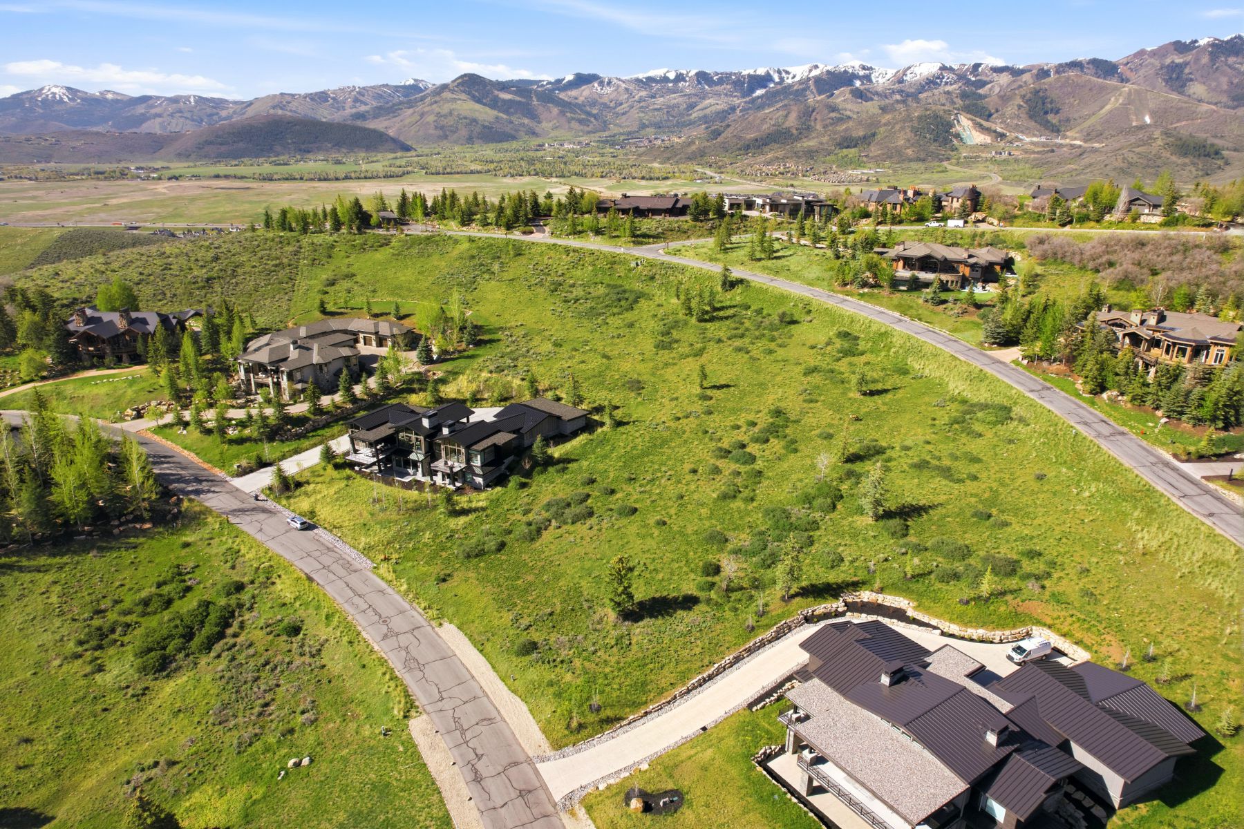 Expansive hillside property featuring a paved driveway, a multi-level residence with a dark metal roof, and distant mountain ranges with snow-capped peaks