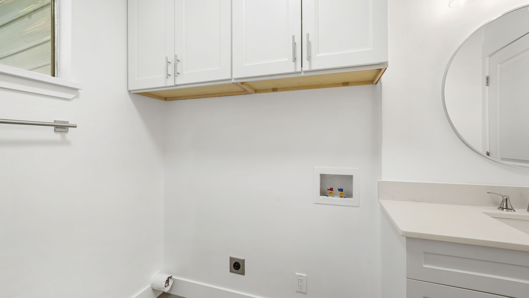 Modern utility space featuring white cabinetry, a built-in vanity with a light-toned countertop, a contemporary circular mirror, and a window providing natural illumination