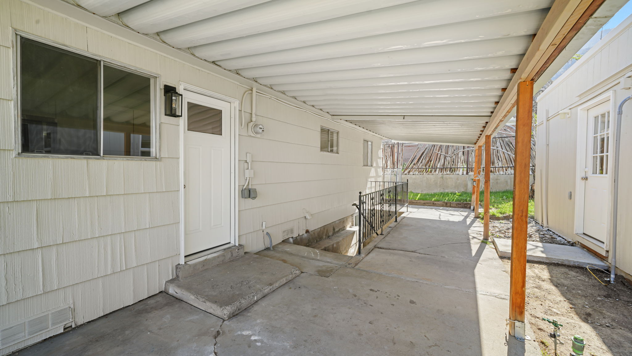 Independent entrance to basement. Covered concrete patio extending the length of the building, featuring a white paneled door with glass insert, black outdoor wall sconce, and white horizontal siding