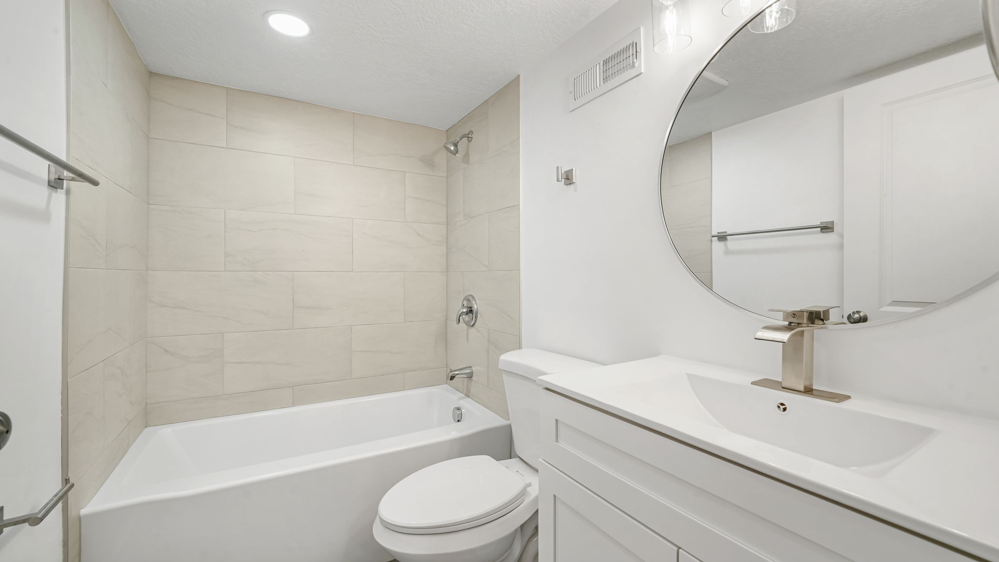 Bathroom featuring a built-in bathtub with light-toned rectangular wall tiling, a white vanity with integrated sink and brushed nickel faucet, a large round mirror, and recessed ceiling lighting