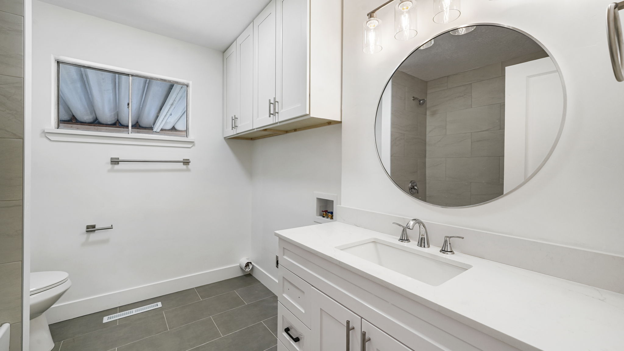 Modern bathroom featuring a single vanity with a white countertop and undermount sink, a circular mirror, and overhead lighting