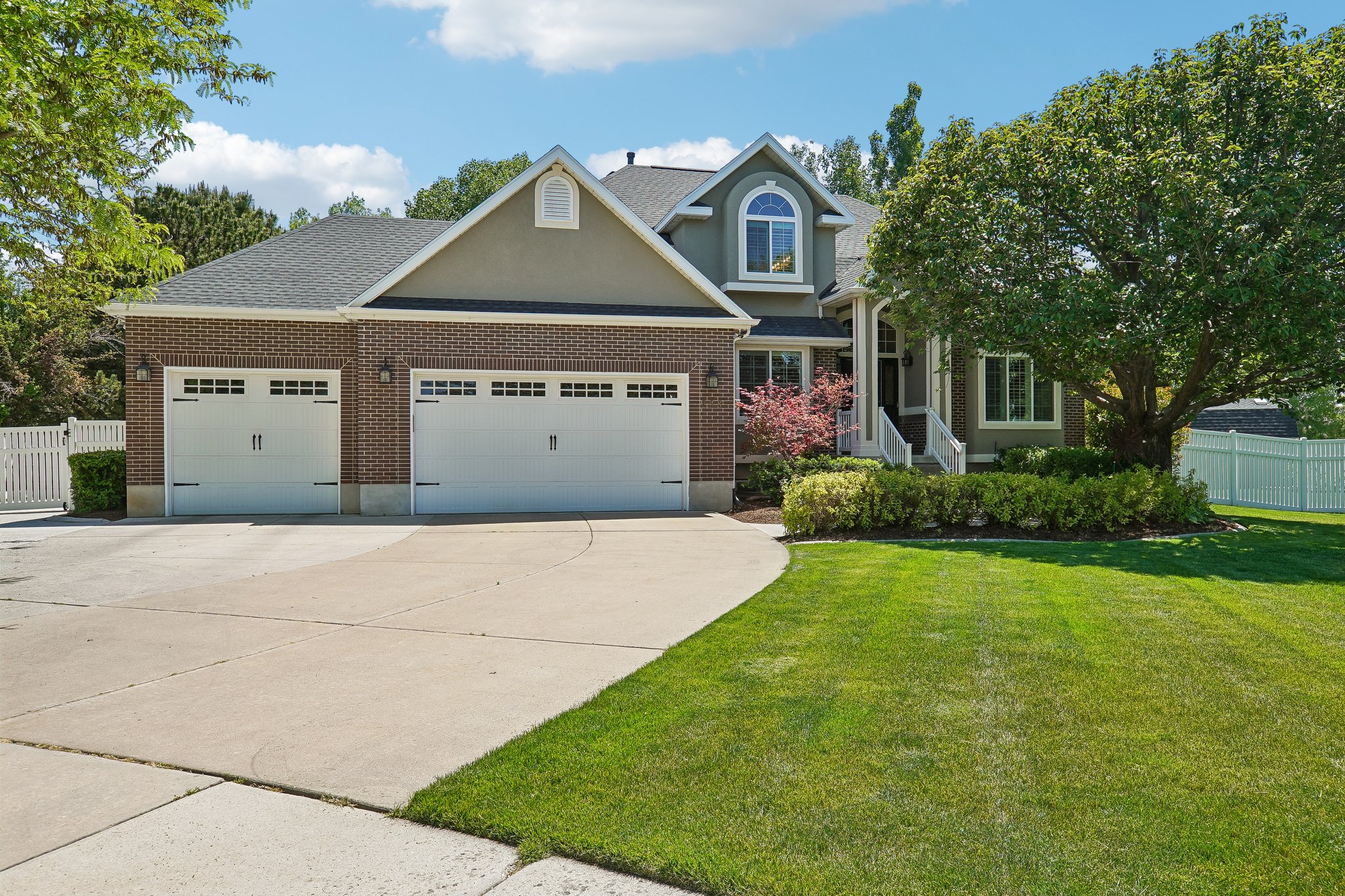 Stately exterior featuring brick and stucco finishes, a multi-car garage, mature landscaping, a white picket fence, and a concrete driveway