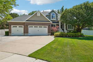 Stately exterior featuring brick and stucco finishes, a multi-car garage, mature landscaping, a white picket fence, and a concrete driveway