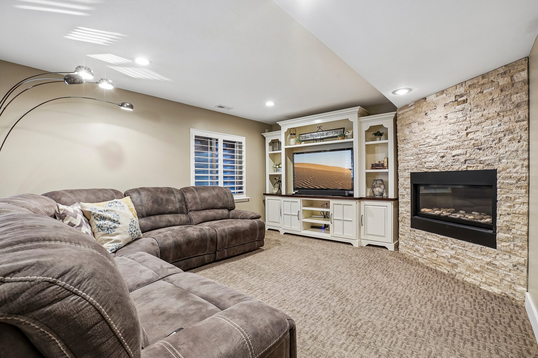 Recessed lighting illuminates a carpeted living space featuring a built-in media center with shelving and an integrated fireplace with a stacked stone surround
