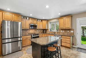 Kitchen featuring a central island with a granite-style countertop, stainless steel appliances, wood cabinetry, tile flooring, and recessed lighting