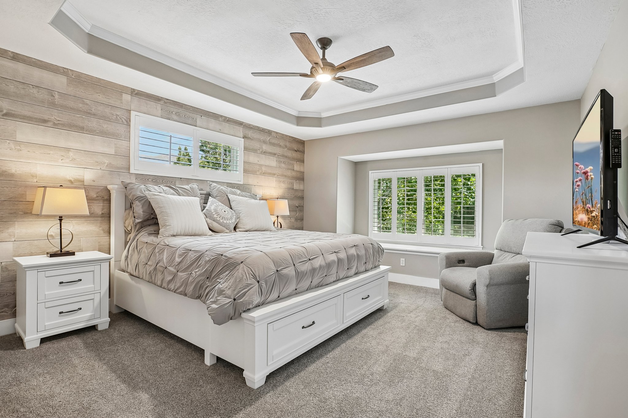 Bedroom featuring a tray ceiling with a ceiling fan, a wood-finish accent wall, and a large window with plantation shutters
