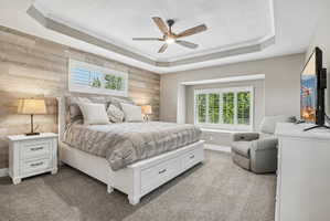 Bedroom featuring a tray ceiling with a ceiling fan, a wood-finish accent wall, and a large window with plantation shutters