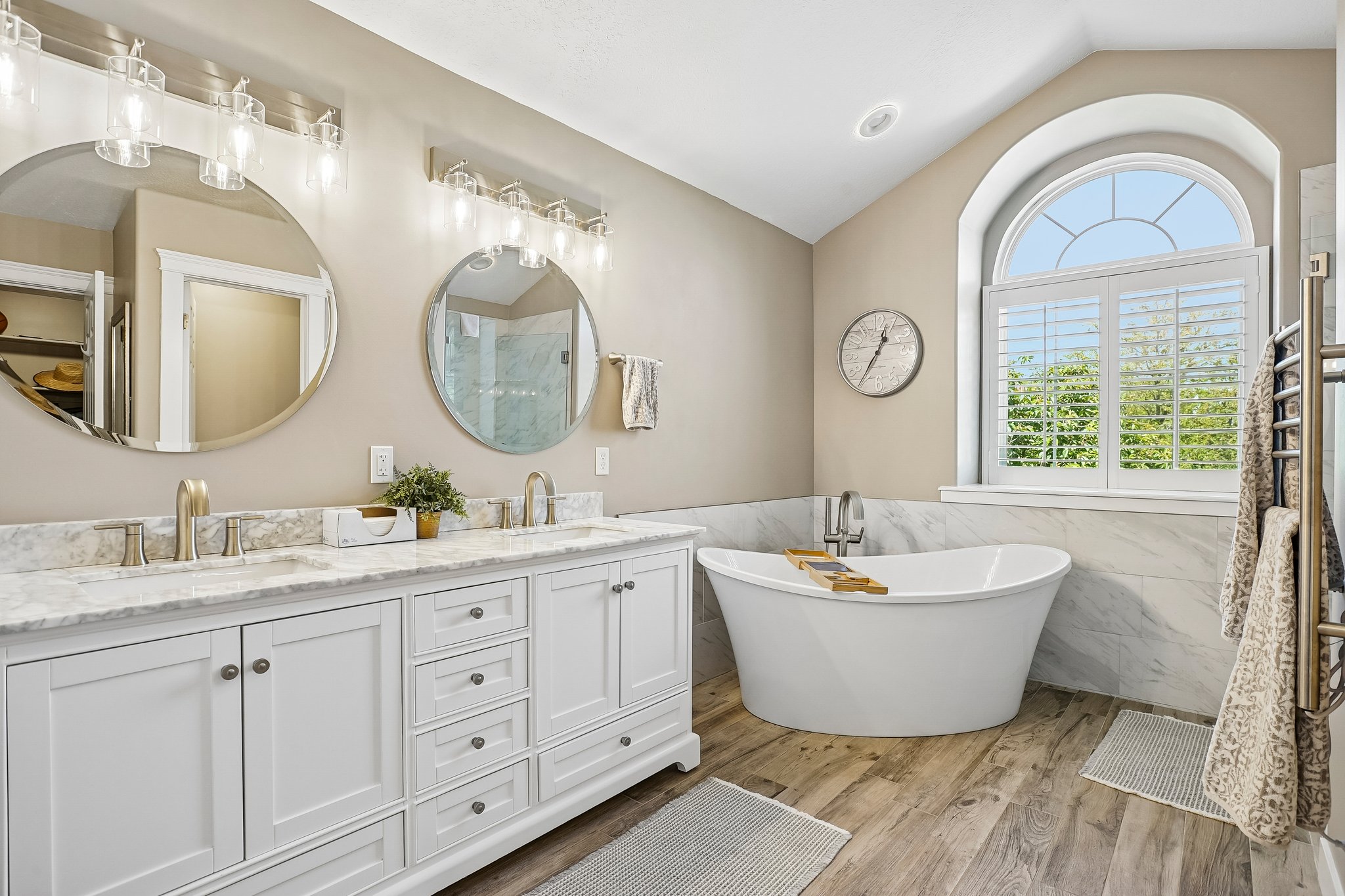 Contemporary bathroom featuring a freestanding soaking tub, arched window with plantation shutters, dual vanity with white cabinetry, stone-finish countertops, and wood-finish flooring