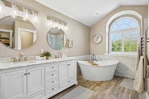 Contemporary bathroom featuring a freestanding soaking tub, arched window with plantation shutters, dual vanity with white cabinetry, stone-finish countertops, and wood-finish flooring