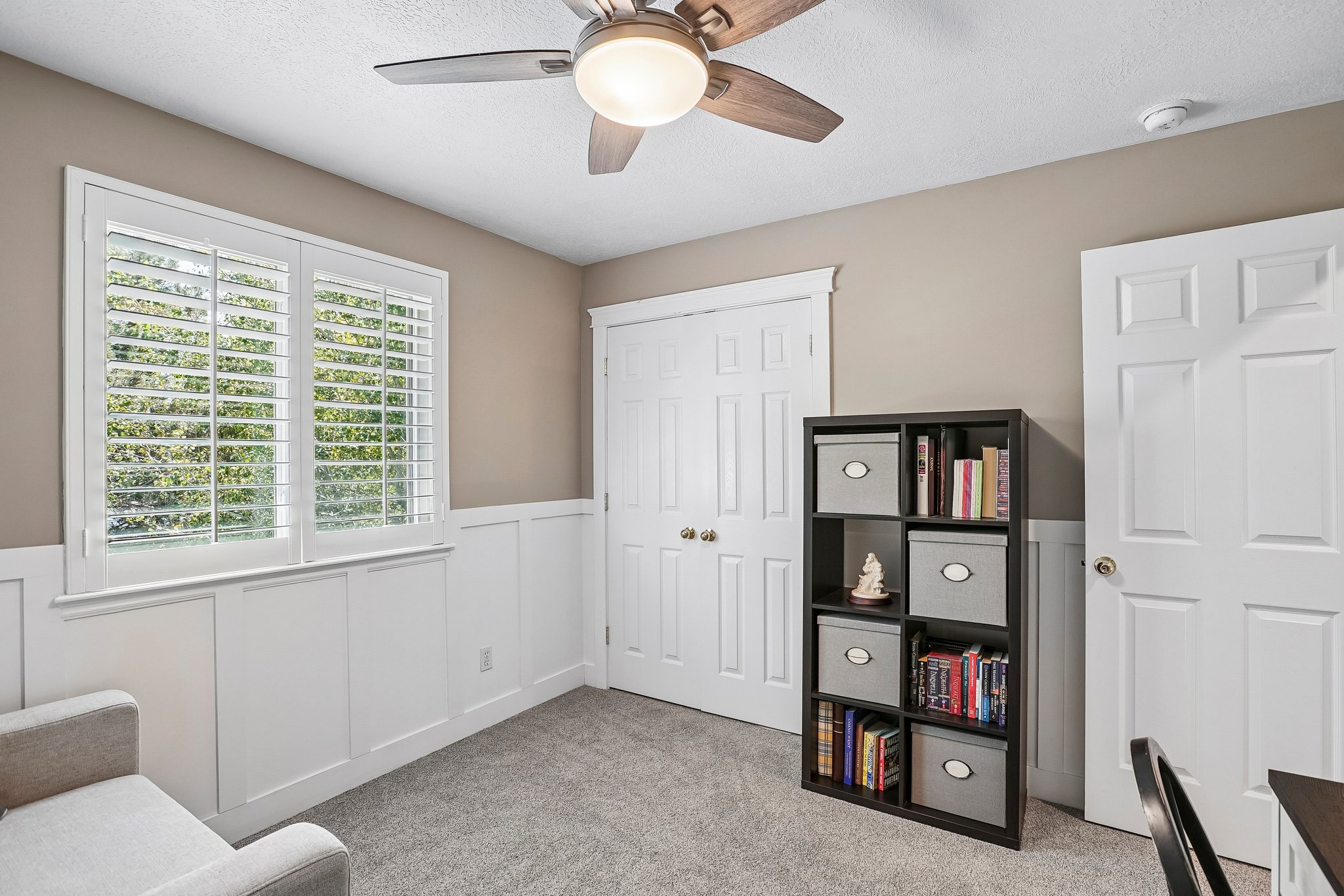 Carpeted room featuring a ceiling fan with light, white wainscoting, and a window with plantation shutters
