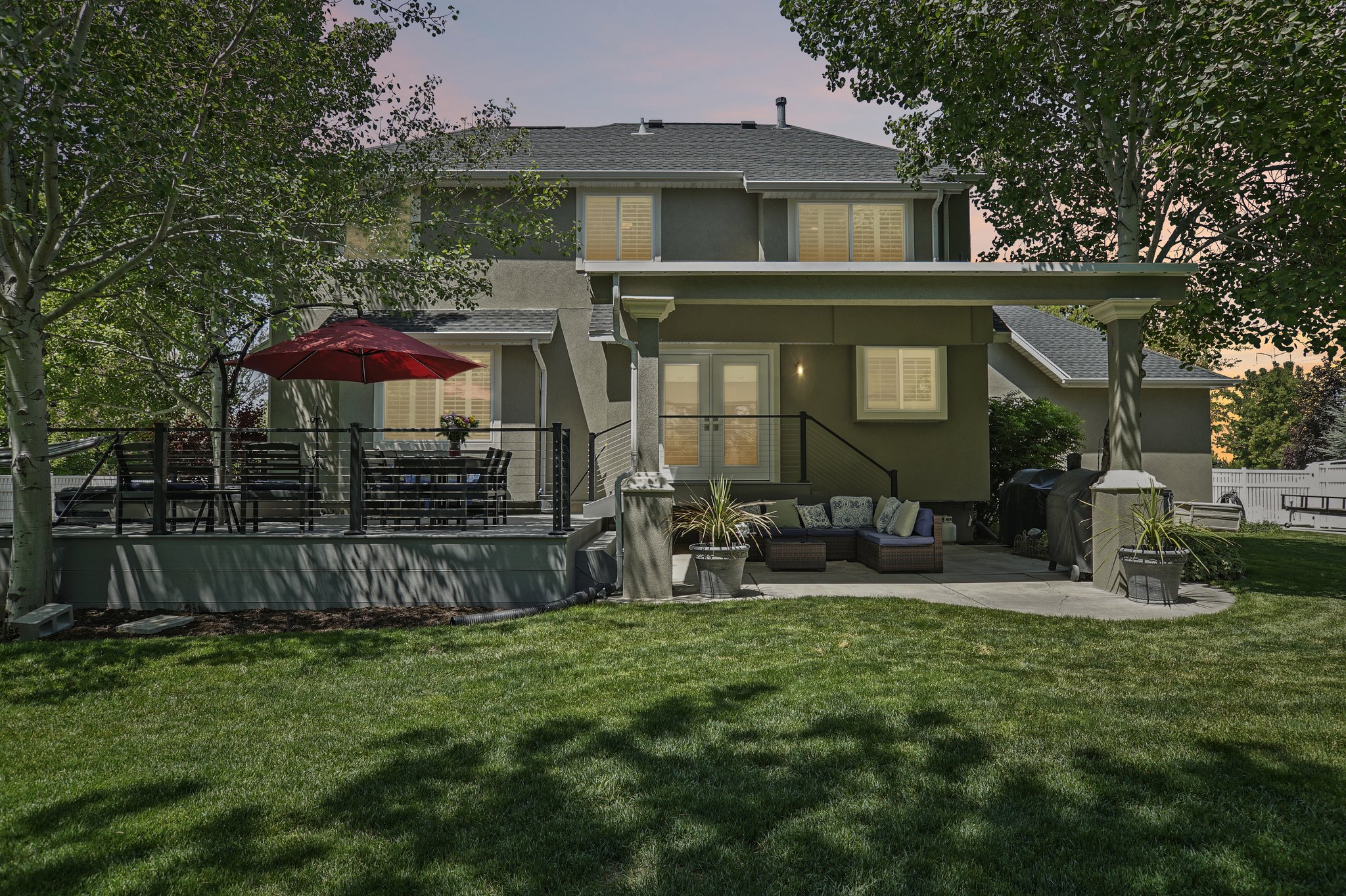 Expansive backyard featuring a manicured lawn, a raised outdoor deck with cable railing, a covered patio area, and mature trees providing shade