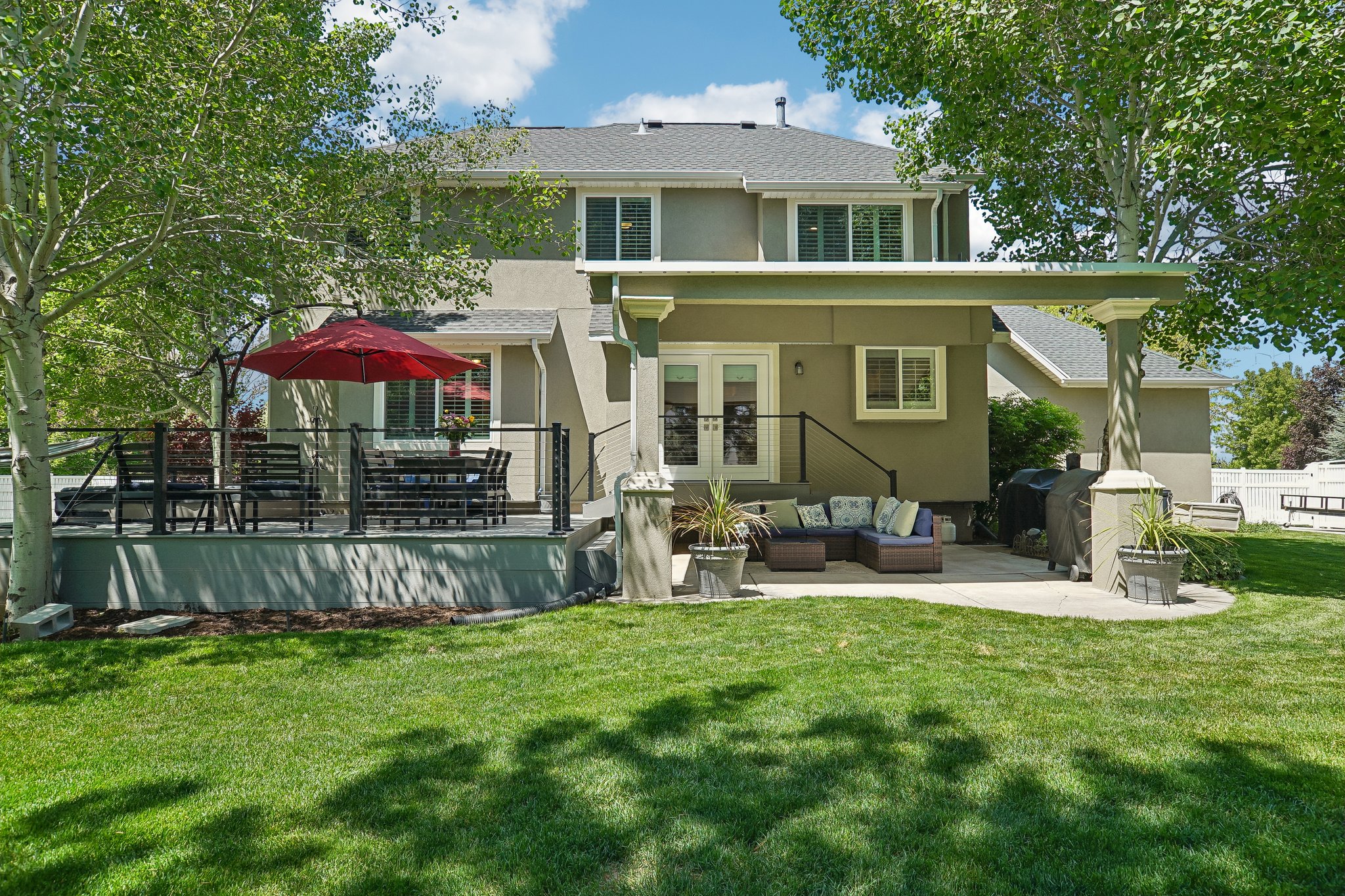 Expansive backyard featuring a manicured lawn, a raised composite deck with cable railing, a ground-level concrete patio, and mature trees providing shade