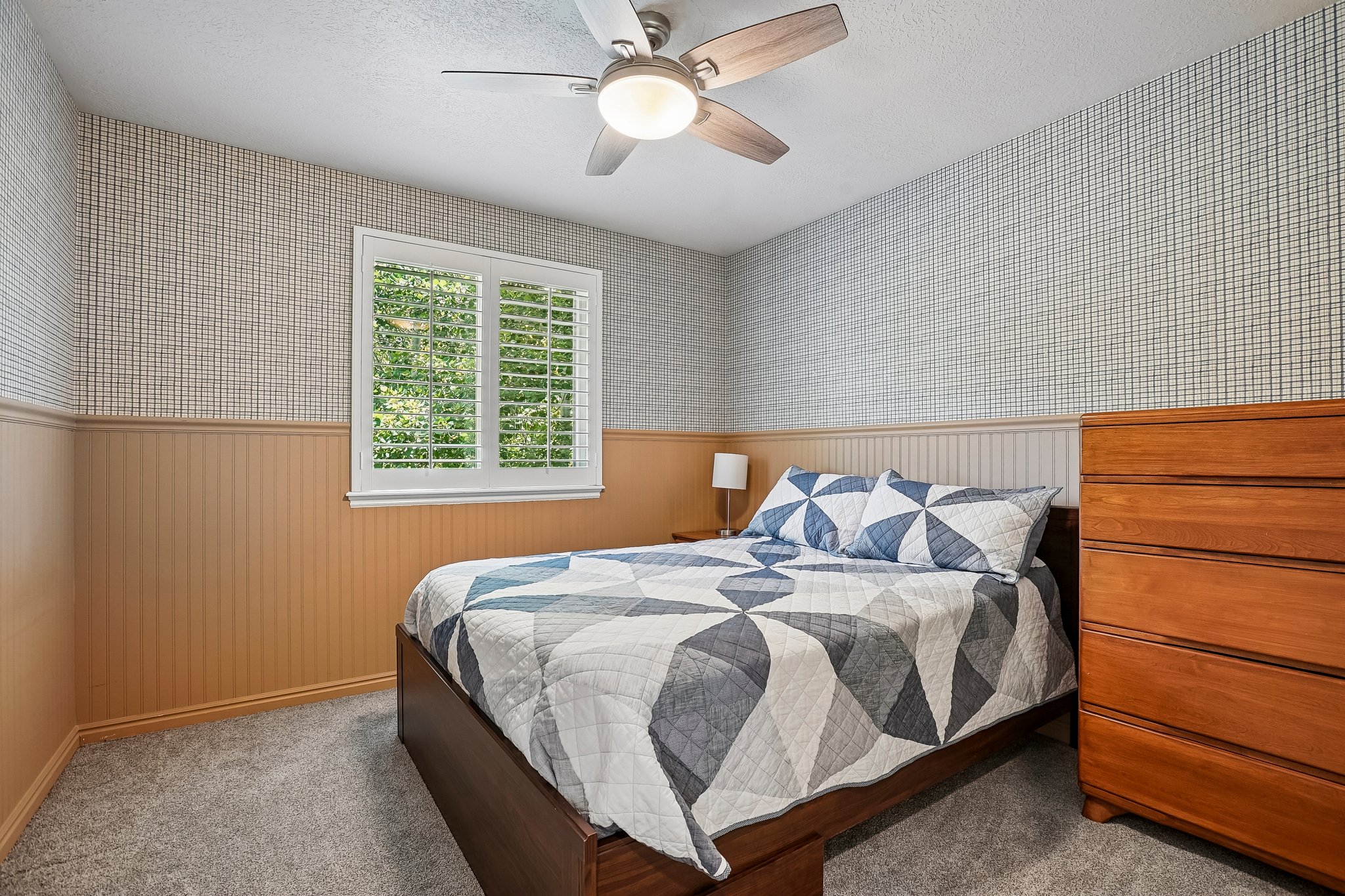 Carpeted interior space featuring a window with white louvered shutters, beadboard wainscoting, patterned wallpaper, and a ceiling fan with integrated lighting