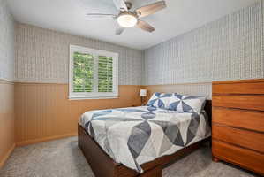Carpeted interior space featuring a window with white louvered shutters, beadboard wainscoting, patterned wallpaper, and a ceiling fan with integrated lighting