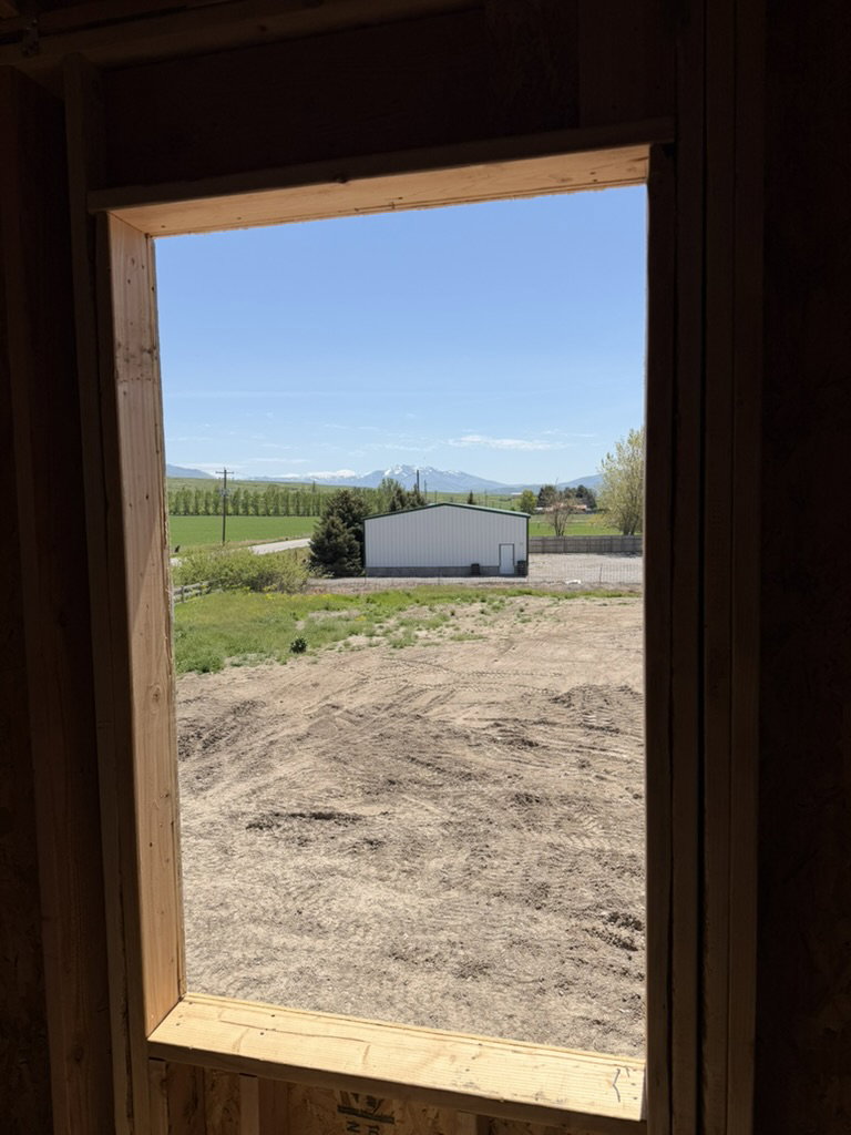 Expansive framed window overlooking a cleared property area, distant agricultural fields, a white metal building, and mountains under a clear blue sky