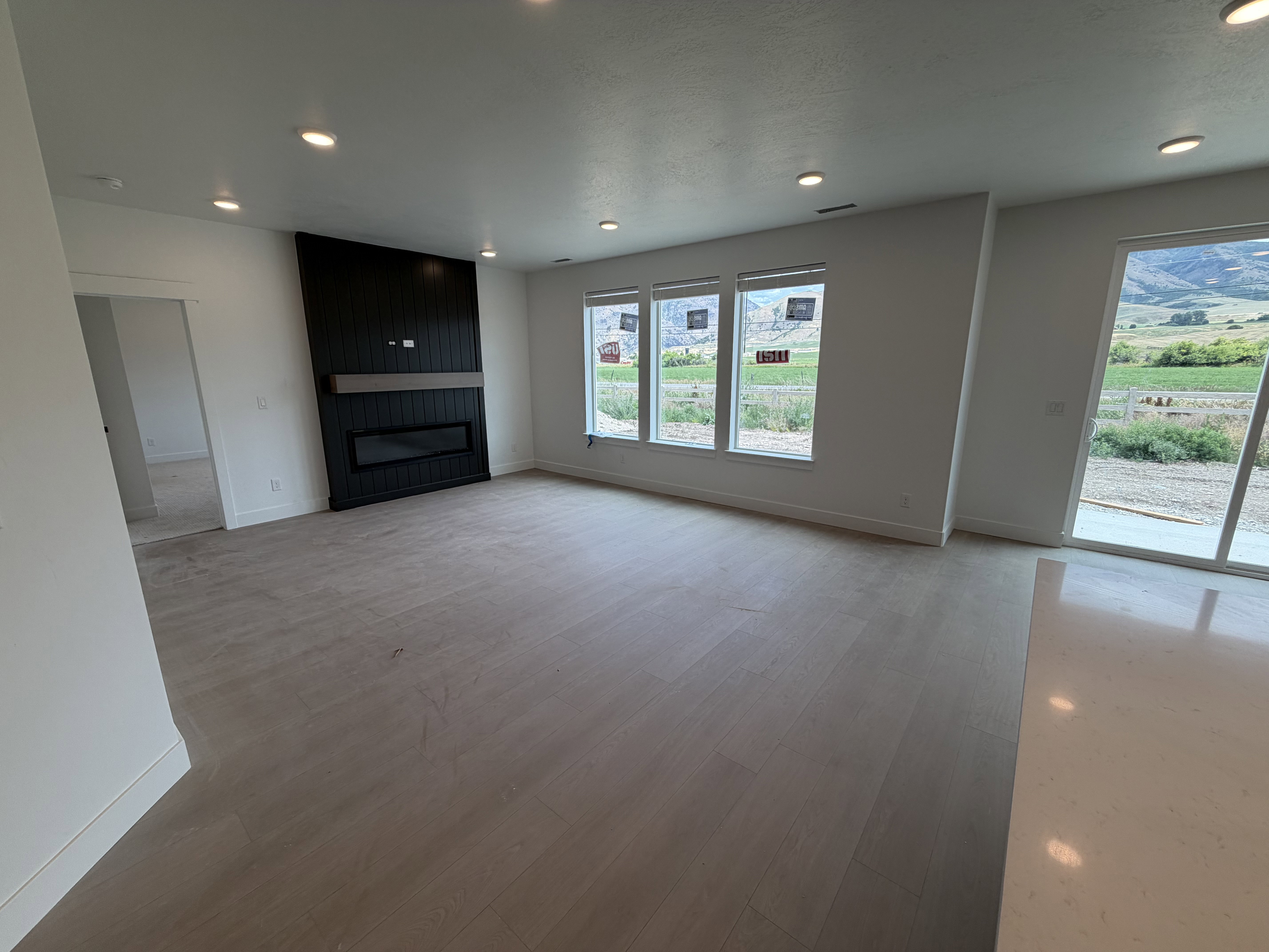 Spacious room featuring light wood-finish flooring, a contemporary fireplace with a sleek black surround and mantel, multiple recessed ceiling lights, a series of three large windows, and an expansive sliding glass door