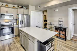Modern kitchen featuring a large central island with a light-toned countertop, stainless steel appliances, neutral cabinetry, and wood-finish flooring