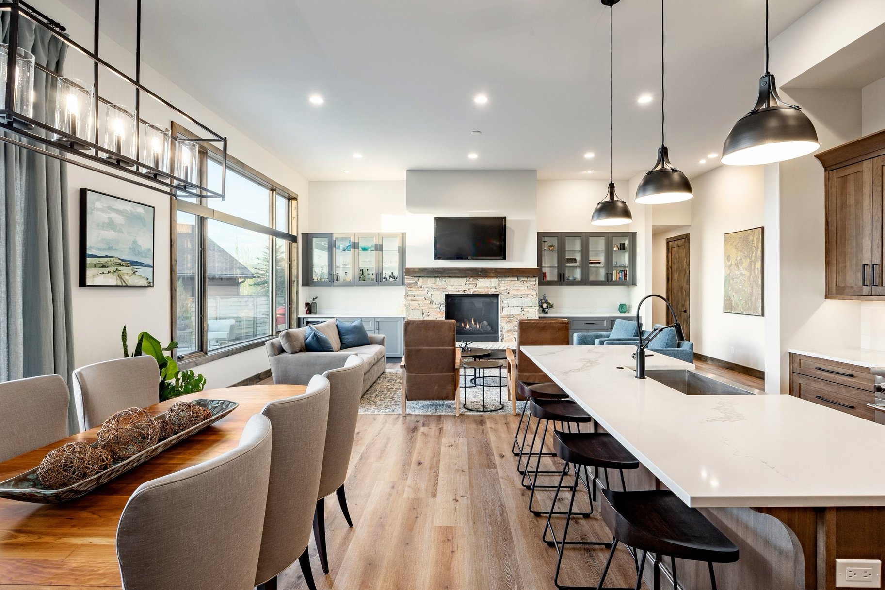 Open-concept interior featuring wood-finish flooring, a stone-clad fireplace, a kitchen island with a built-in sink, pendant lighting, and expansive windows