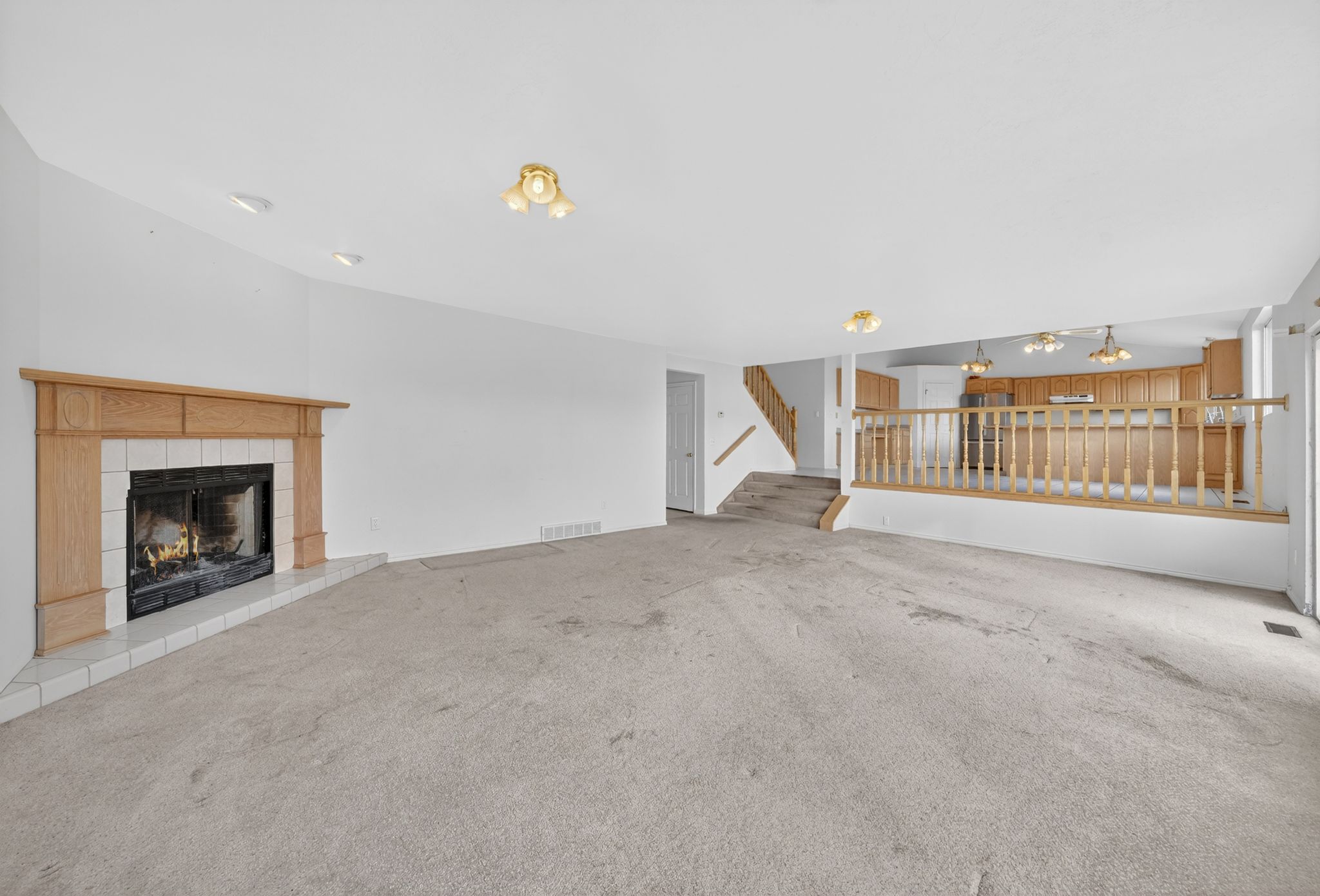 Carpeted living area featuring a fireplace with a wood mantle and tile surround, recessed lighting, and a half-wall with wooden railing overlooking a kitchen area with wood cabinetry