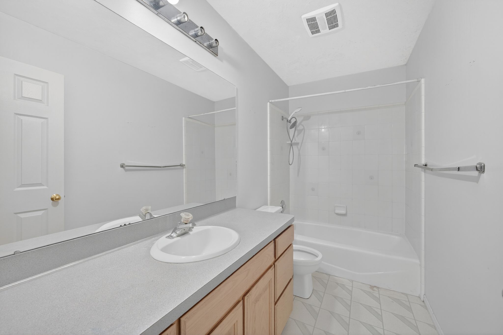 Bathroom featuring a light wood-finish vanity with a single basin sink, a gray countertop, an expansive wall-mounted mirror, a tub/shower combination with a clear enclosure, and patterned floor tiling