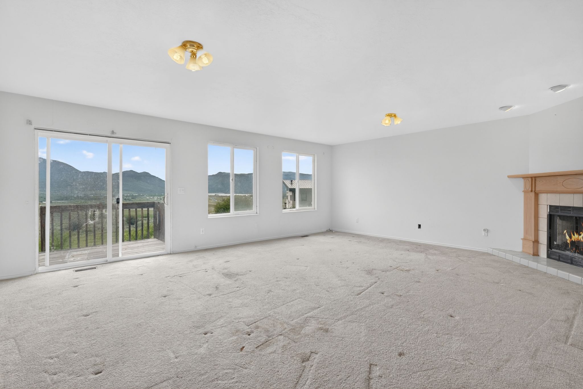 Spacious living area featuring a fireplace with a wood mantle and tile surround, light-colored carpeting, multiple windows, and a sliding glass door opening to an outdoor deck with mountain scenery