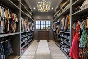 Custom closet featuring extensive built-in shelving, hanging rods, an ornate chandelier, a window with exterior scenery, and a central upholstered bench