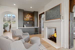 Living space featuring a stone fireplace with a herringbone tile interior, a built-in wet bar with a wine refrigerator, light wood-finish flooring, an arched doorway, and exposed wood beams