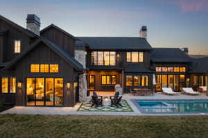 Expansive rear exterior featuring a swimming pool with integrated spa, stone patio, dark wood siding, multiple large windows, and prominent stone chimneys