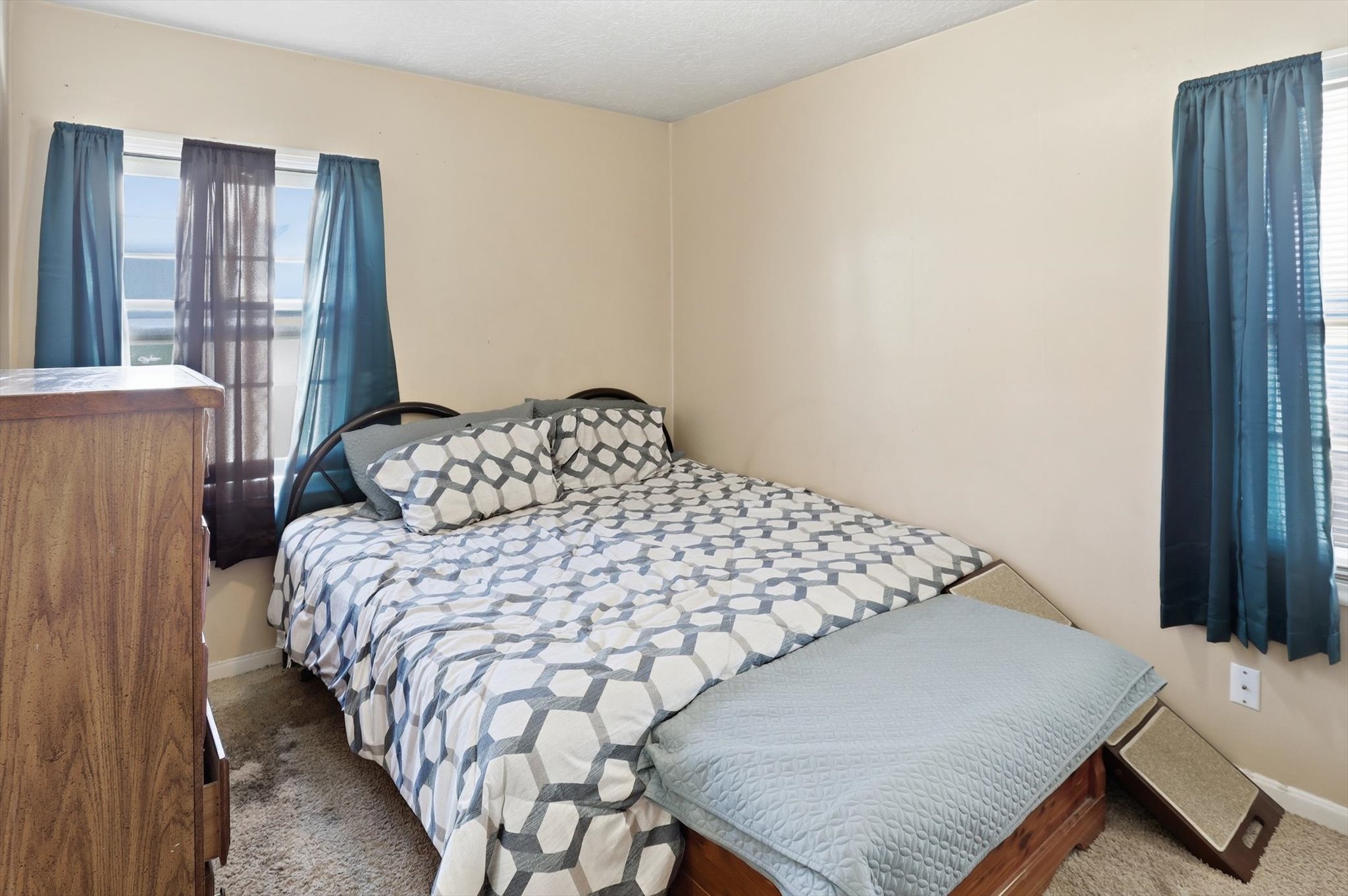 Bedroom featuring two windows with blinds, neutral wall paint, and textured carpeting