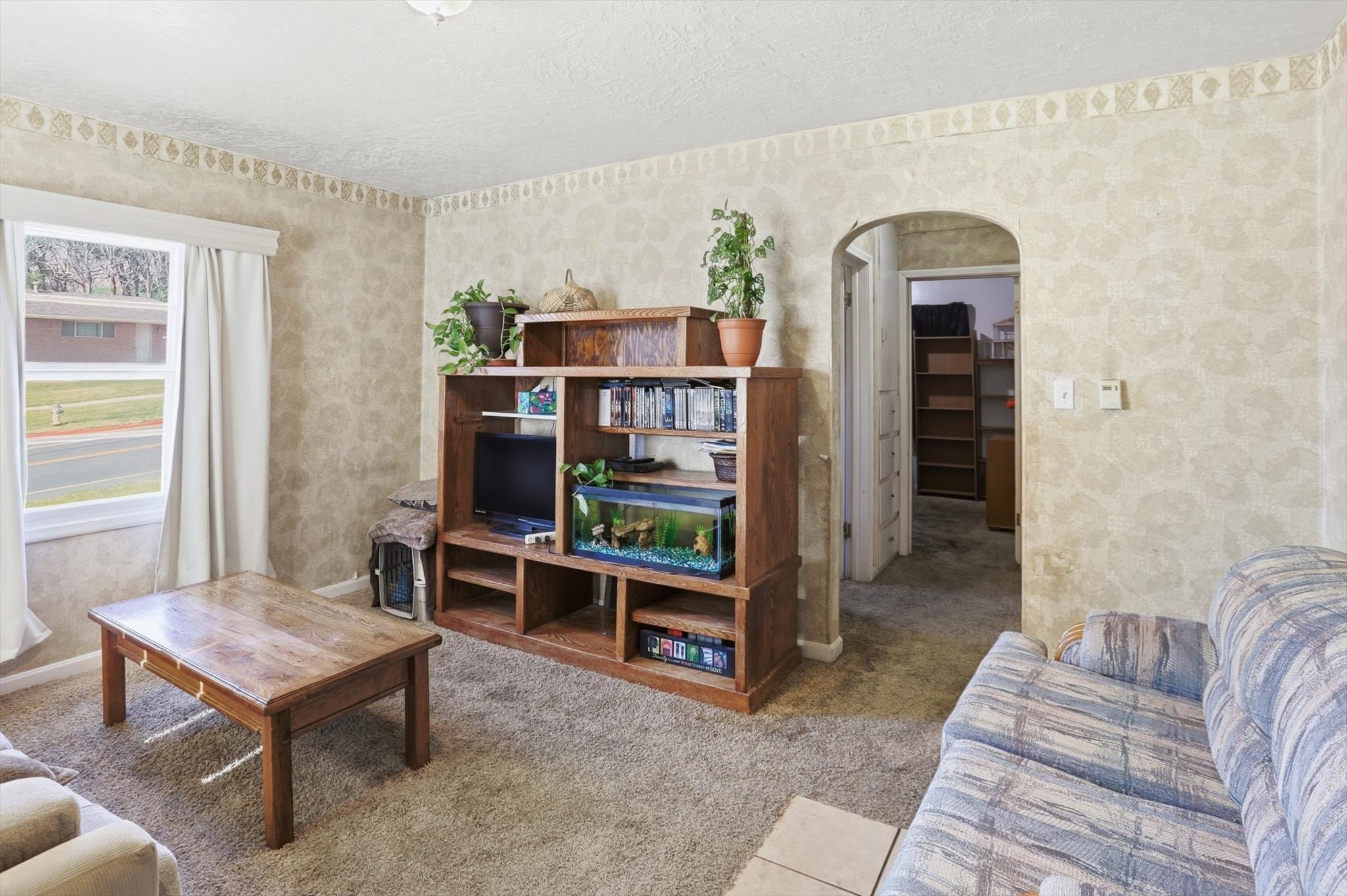 Carpeted living area featuring a large window with white curtain panels, textured wallpaper, and an arched doorway