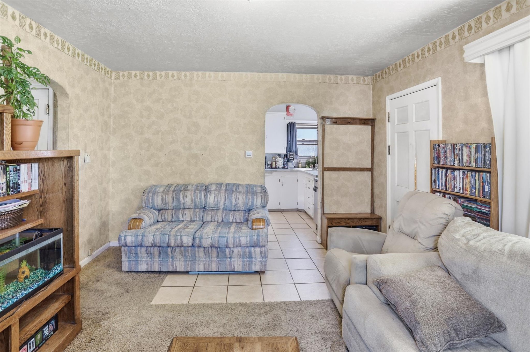 Living area featuring carpet flooring, patterned wall coverings, a two-panel interior door, and an arched entryway leading to a tiled-floor space