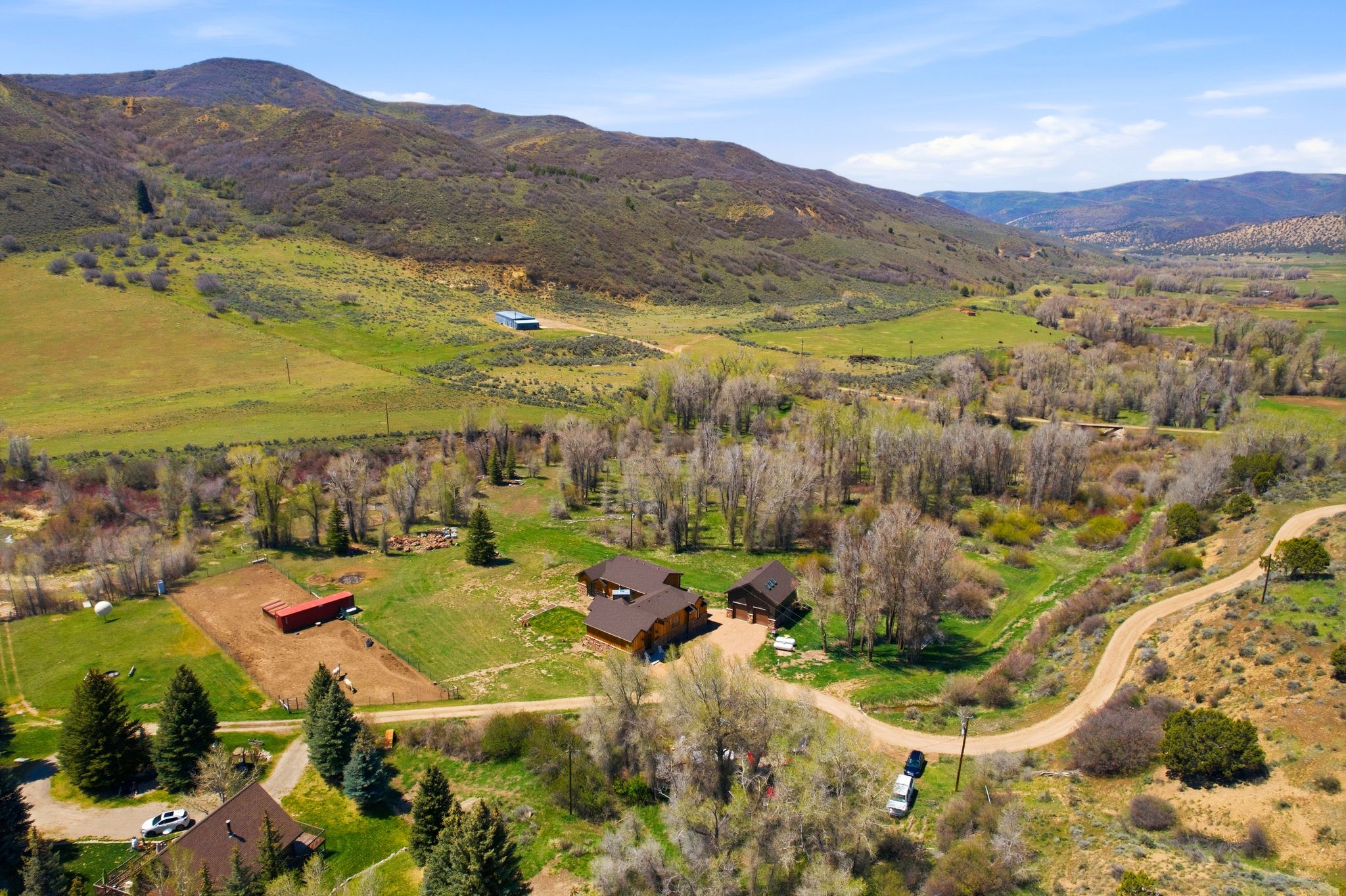 Expansive mountain valley property featuring multiple structures, verdant fields, a dirt road, and abundant mature trees