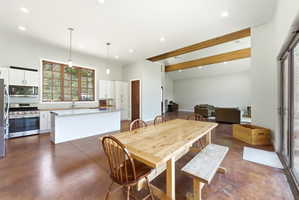 Open-concept living space with stained concrete flooring, exposed wood ceiling beams, a kitchen island with seating, stainless steel appliances, and a large multi-pane window