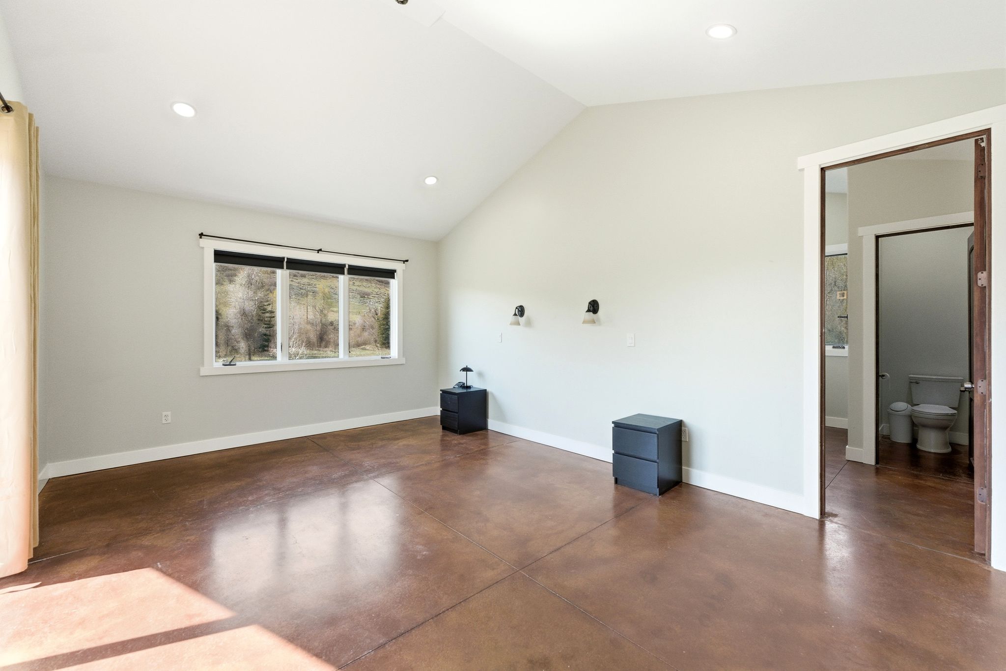 Vaulted ceiling room with stained concrete flooring, recessed lighting, wall-mounted sconces, and a large window overlooking natural scenery