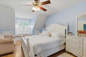 Bedroom featuring warm wood-finish flooring, a ceiling fan with integrated lighting, and a window with white plantation shutters