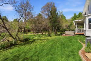Expansive green lawn surrounded by mature trees, featuring a covered wooden deck with white railing, and a hot tub on a brick patio